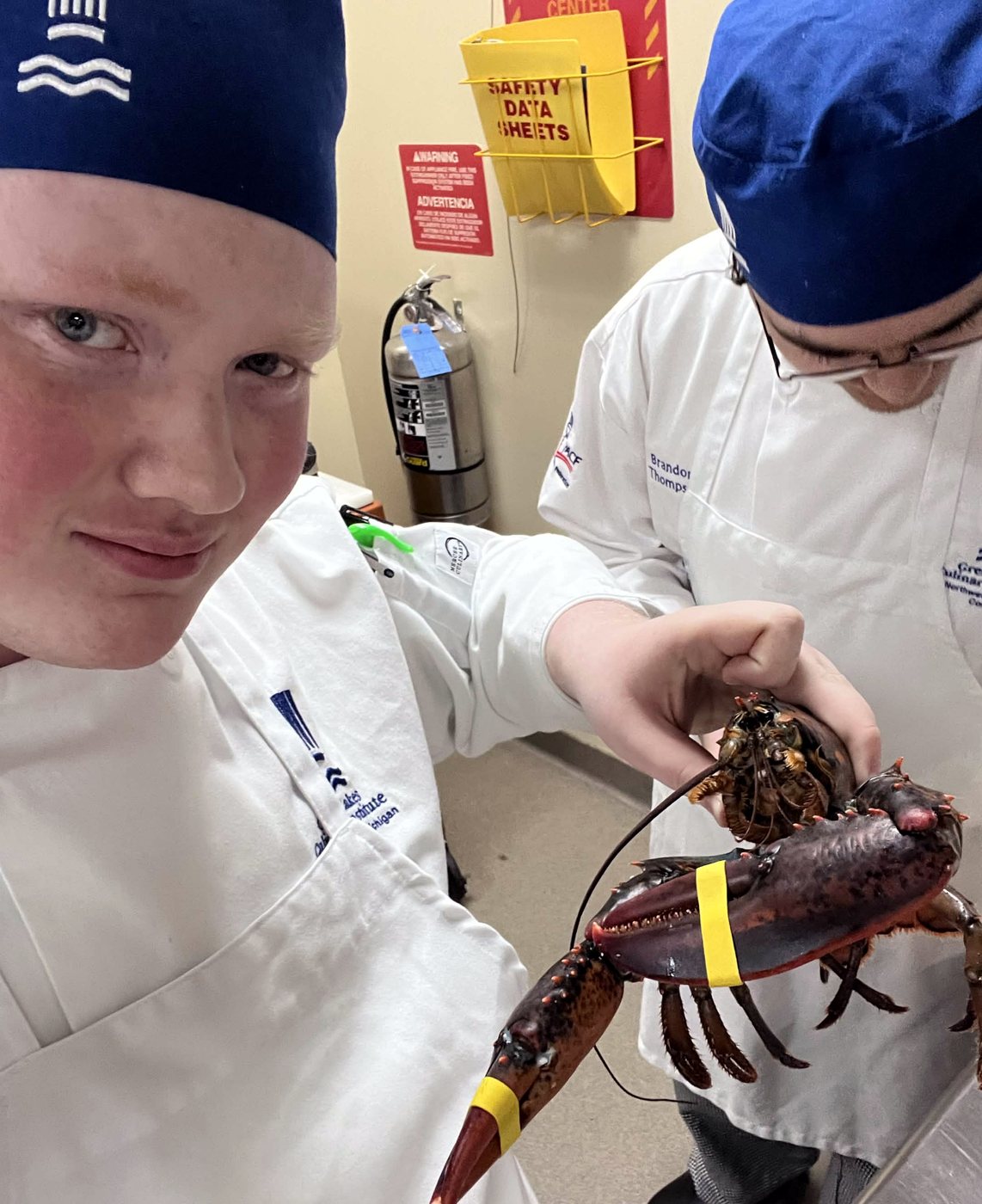 Alex in a chef coat and blue cap holding a live lobster in a teaching kitchen