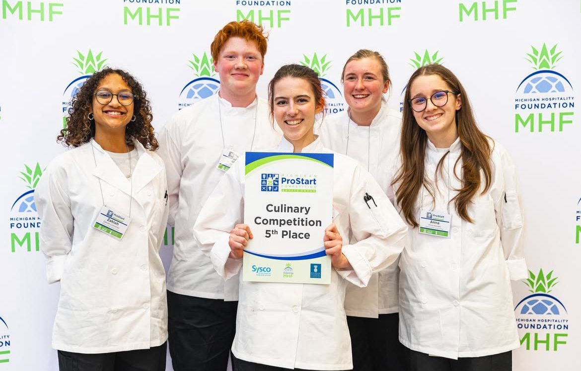 Five students in chef coats holding a fifth place sign in front of a Michigan Hospitality Foundation backdrop