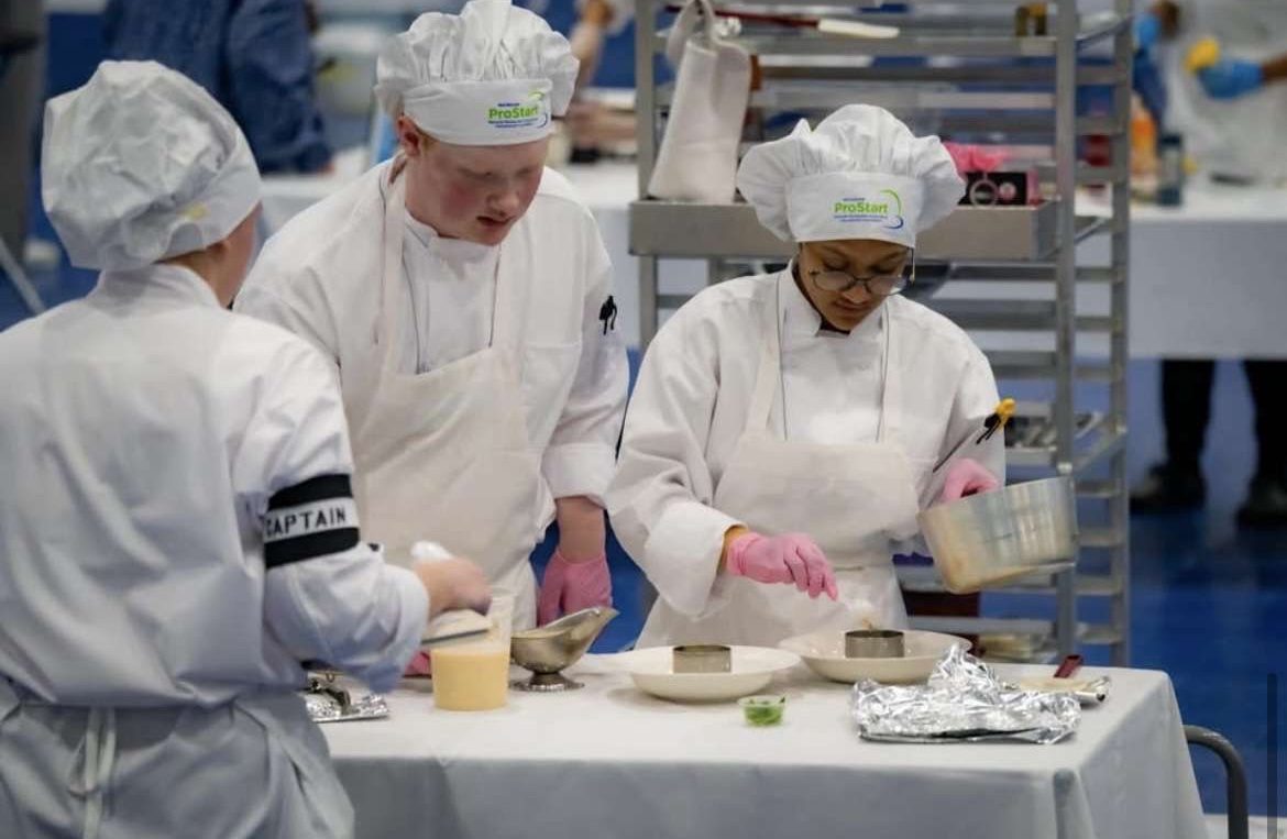 Three students in chef whites plating dishes at a competition table