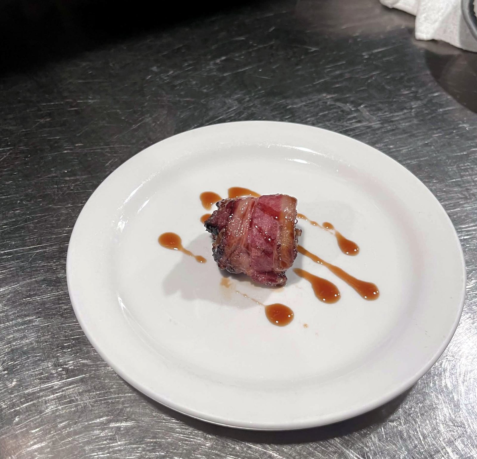 A single bacon wrapped date on a white plate with dots of sauce